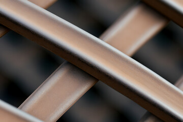 A close-up shot showcases an abstract pattern created by intersecting metallic bars. The subtle interplay of light and shadow emphasizes the texture. The metal is warm toned.
