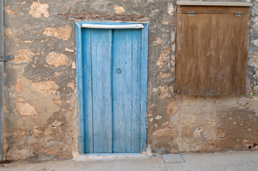 Sunlit blue door on stone facade with boarded hatch, narrow sidewalk leading past shuttered entrance, aged stucco and rough texture, quiet travel photography mood