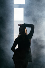 A mysterious woman stands in silhouette against a bright window, surrounded by dramatic smoke and shadows