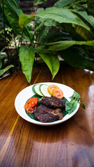 A traditional Indonesian dish of fried meat, possibly empal, served with spicy sambal, cucumber, and tomato on a wooden table with a lush plant background.