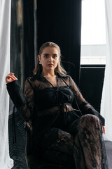 Confident woman in sheer black lace stands poised between white curtains, embodying strength, elegance, and modern gothic fashion