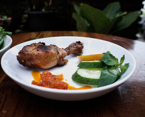 A close-up shot of a delicious chicken leg served with fresh cucumber slices and a side of spicy sauce on a white plate.