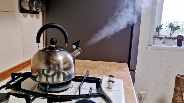Side view of a stainless steel kettle boiling on a gas stove in a kitchen. A powerful horizontal jet of white steam shoots from the spout as the water boils vigorously over a bright blue flame.