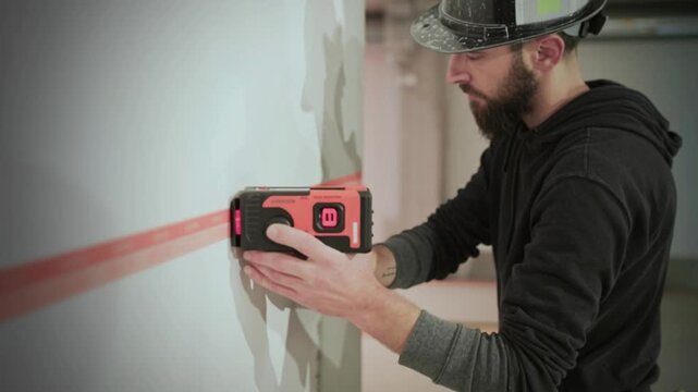 Construction worker scanning interior wall with electronic stud finder laser measuring device, professional renovation and building inspection concept showing precise alignment tool used for accurate 