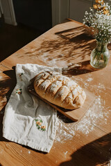 Fresh homemade rustic bread on a light wooden table, linen napkin, scattered flour, a vase of wildflowers, cozy quiet morning, slow living, homemade food, bakery, and rustic lifestyle concepts