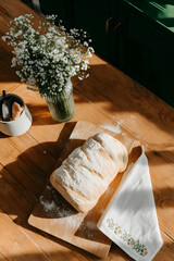 Fresh homemade rustic bread on a light wooden table, linen napkin, scattered flour, a vase of wildflowers, cozy quiet morning, slow living, homemade food, bakery, and rustic lifestyle concepts