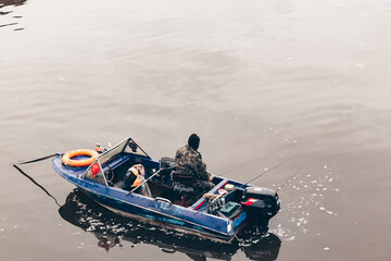 Man fishing from a blue boat in a canal during autumn. A bright lifebuoy rests at the bow, surrounded by calm water and colorful fall reflections