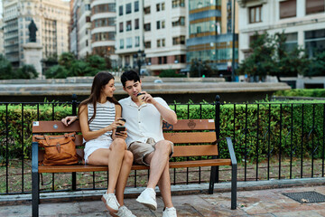 Fototapeta premium Portrait of a happy young couple, woman and man, having fun using a mobile phone and sitting on bench and talking and bonding in park outdoors