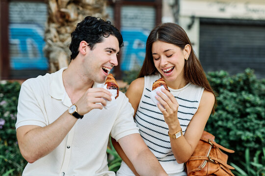 Fototapeta Happy couple  eating a pastry croissant, smiling and looking away on a city street