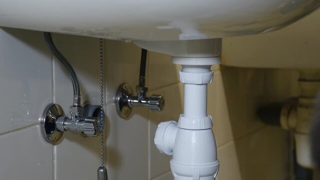 Professional plumber performing maintenance, tightening a plastic bottle trap drain pipe by hand under a white ceramic bathroom sink, ensuring a proper seal and preventing water leaks