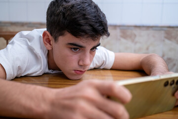 Obraz premium Teenage boy lying on a table, intensely focused on his smartphone screen, in a quiet indoor environment, wearing a white t-shirt.