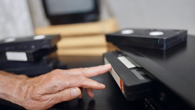 Senior male's hand puts the tape into VHS recorder. Close up. A pile of tapes and an old screen at blurred backdrop.