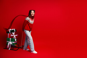 Young woman in red sweater pulls a sled with gifts while smiling against a bright red background capturing festive Christmas joy and holiday spirit