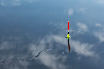 Red yellow and black fishing bobber float standing vertically in calm blue water with cloud reflections and ripples. Serene fishing scene showing tackle equipment in peaceful lake.