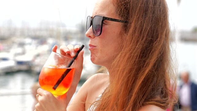Woman enjoying refreshing orange cocktail drink at scenic marina waterfront location with sailing boats Gijon Spain on summer day