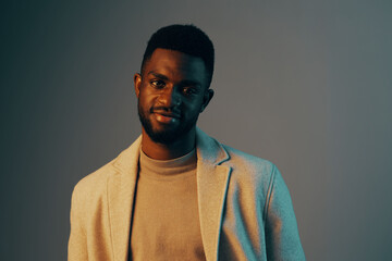 Obraz premium A stylish young African man wearing a beige blazer and a light sweater smiles confidently against a soft blue backdrop, exuding modern elegance and charm