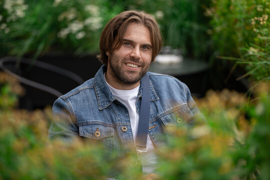 A smiling man with a beard and a denim jacket is seen through green foliage.