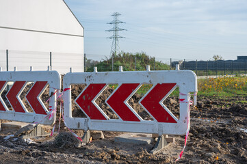 Road construction site with red and white chevron barricades blocking muddy ground near building...