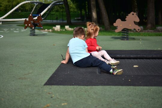 Two little children sitting together on trampoline border outdoors at playground, relaxing after active play, cute peaceful childhood moment in park nature environment.