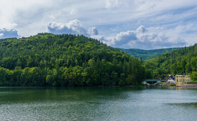 Serene mountain lake bordered by dense green forest and soft rolling hills under cloudy summer sky. The tranquil water surface reflects lush vegetation, creating a calm natural landscape ideal for tra