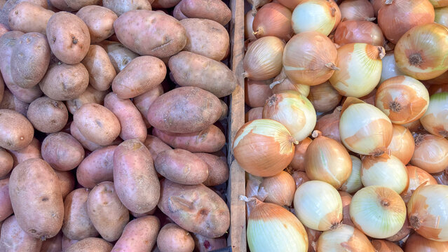 Asterix potatoes (reddish-pink) and yellow onions displayed in bulk. Fresh vegetables on display for sale at a market or fair, separated by a dark divider.