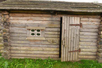 The facade of a traditional log structure (a barn or shed) is built from horizontal logs, featuring a small window and an open plank door, rising from green grass