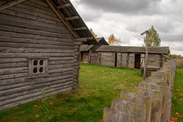 Weathered log houses and utility buildings of a Slavic settlement reconstruction stand behind a simple wooden fence, featuring traditional architecture under a cloudy sky
