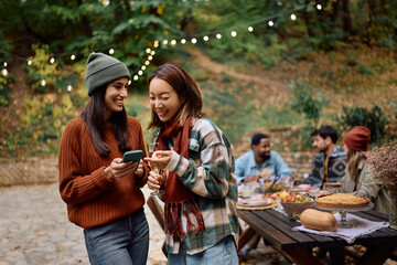 Young happy women using cell phone while gathering with friends on Thanksgiving on patio.