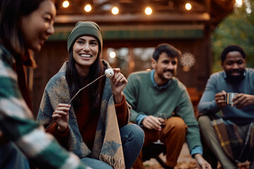 Happy woman and her friends roasting marshmallows in backyard in autumn.