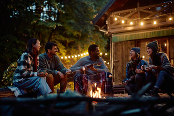 Multiracial group of happy friends having fun while gathering around campfire in autumn.