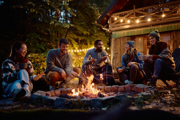 Group of happy friends talking while roasting marshmallows over campfire on autumn evening.