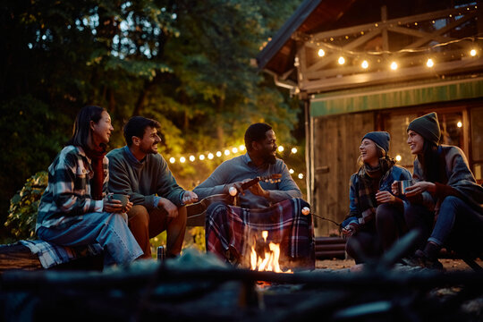 Multiracial group of happy friends having fun while gathering around campfire in autumn.
