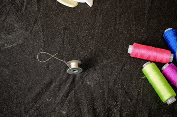 sewing supplies on dark fabric background with colorful spools of thread and a metal bobbin with a piece of thread, concept of needlework, handmade crafts, sewing accessories, and creative hobby.