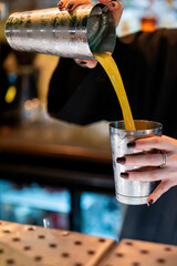 A bartender pours a vibrant, yellow cocktail from a metal shaker into a glass, captured in a close-up shot that highlights the action in a bar setting.