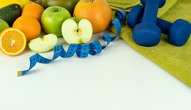 The image shows fresh fruits (oranges, grapefruit, avocado, and green apples, one of which is cut), a blue measuring tape, and two blue dumbbells on a green towel, all of which symbolize a healthy