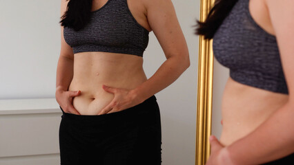 Woman examining her stomach in front of a mirror, gently holding belly skin. Concept of body awareness, self-acceptance, realistic beauty, fitness progress, and confidence in everyday body image.