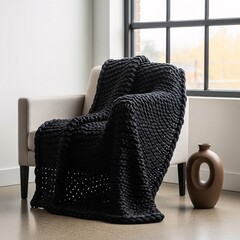 Modern home interior with a cozy black chunky knit blanket draped over a beige armchair by a large window. Hygge concept for stylish, comfortable living room decor and relaxation during autumn