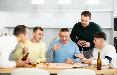 Fototapeta premium Man drinking shot while losing fun alcohol-fueled board game surrounded by laughing friends during friendly get-together at home kitchen..