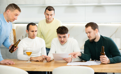 Fototapeta premium Upset worried man sitting at table in home kitchen with papers in hands, reading unexpected medical results with group of male friends solacing and comforting him ....