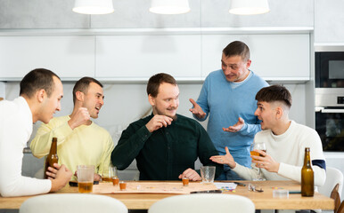 Defeated man taking shot of liquor surrounded by carefree excited male friends playing fun drinking board game during bachelor party at home