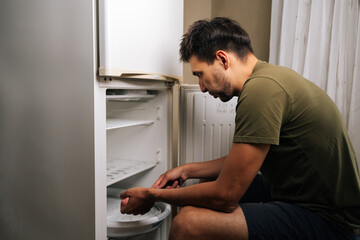 Side view of man sitting on stool, defrosting refrigerator and cleaning interior, performing essential maintenance in kitchen, ensuring hygiene and efficiency in daily routines.