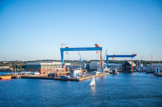 German Naval Yards, Kiel, Germany, sailboat glides through a bustling harbor under a clear blue sky, with large cranes and industrial buildings in the background