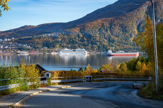 A scenic view of Narvik, Norway harbor with Aida Bella cruise ship and cargo ship, set against a backdrop of autumnal mountains and a curving road