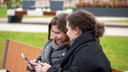 Two Friends Enjoying a Moment Together While Checking Their Phones on a Park Bench in Autumn