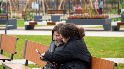 Friends Enjoying Time Together on a Park Bench While Using a Smartphone in a Vibrant Outdoor Setting