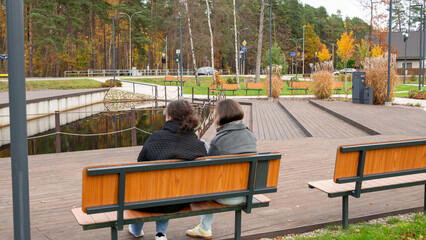Two Women Sit on a Bench by the Water in a Park During Autumn in a Quiet Neighborhood