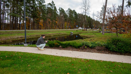 Person Kneeling by a Small Pond in a Park Surrounded by Trees During a Cloudy Autumn Day