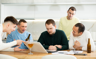 Group of men gathered for friendly meeting with beer at cozy home kitchen, trying to work through challenging problem, viewing laptop screen with concerned expressions and gestures ..