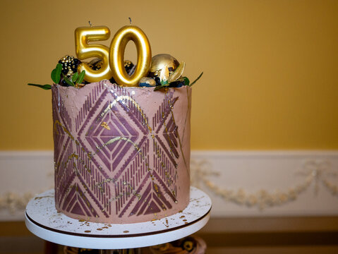 A pink cake with a gold geometric pattern and the number "50" on top is displayed on a white stand against a yellow wall