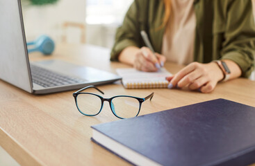 Close-up of eyeglasses on a table with a woman writing in a notepad in the background. Business...
